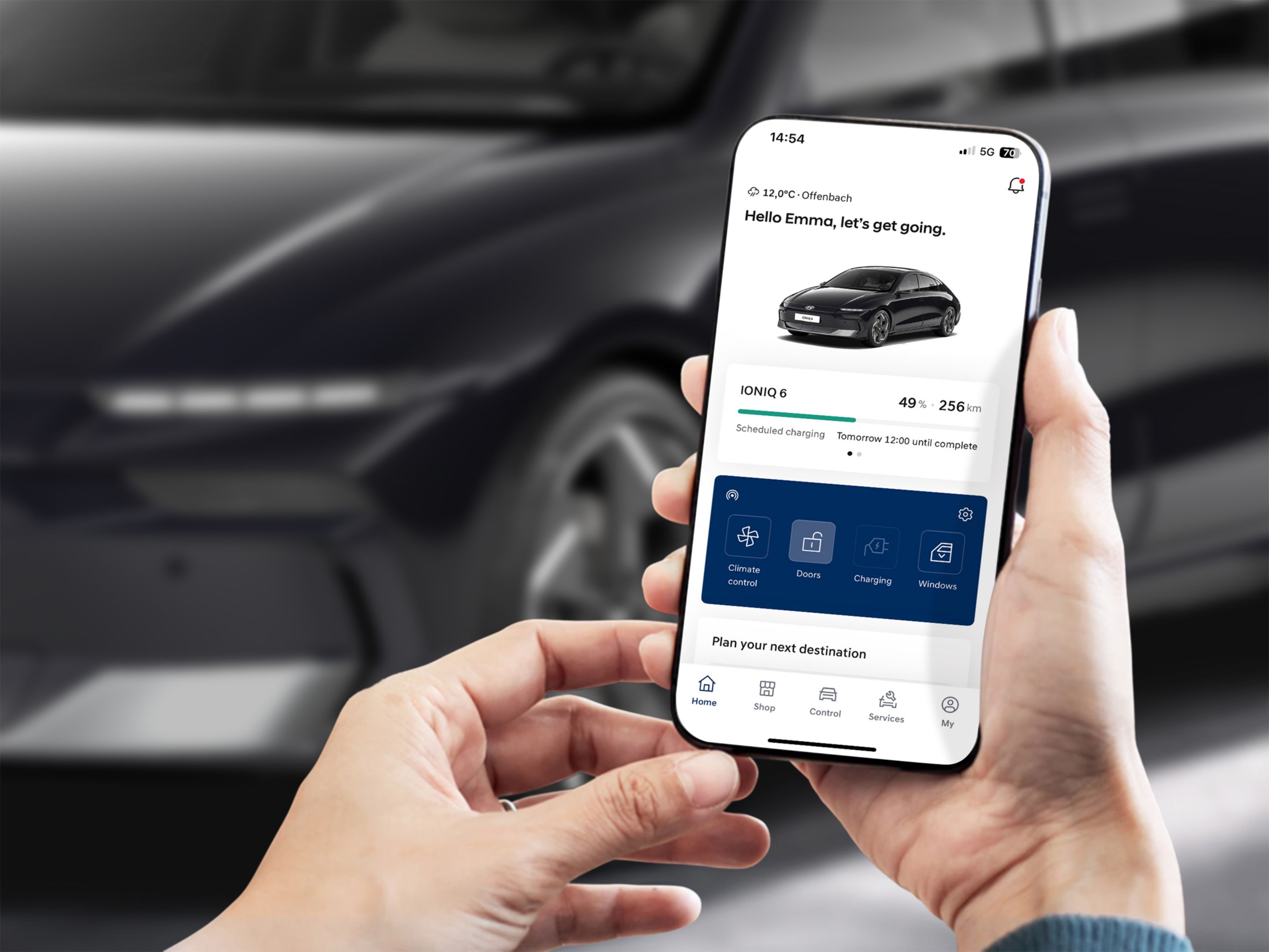 A person is looking at a smartphone with myHyundai app. In background is Hyundai IONIQ 6 parked on the street.