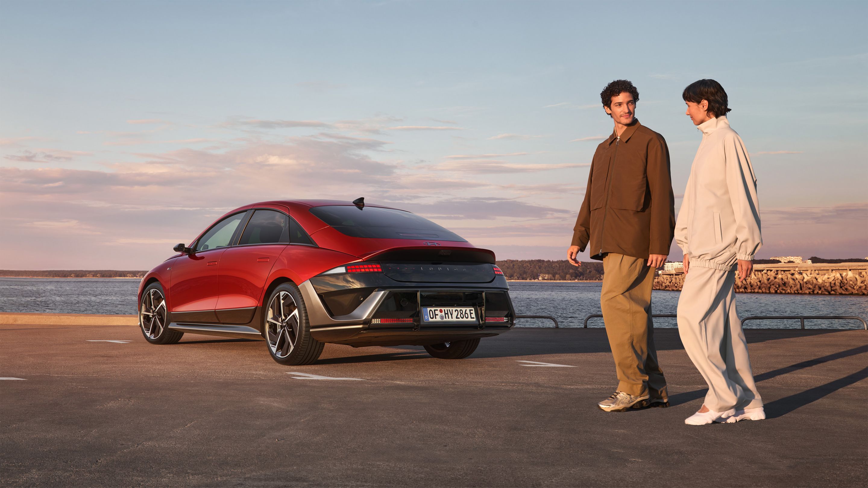 A man and a woman walking past a red Hyundai IONIQ 6 N Line parked next to the ocean.