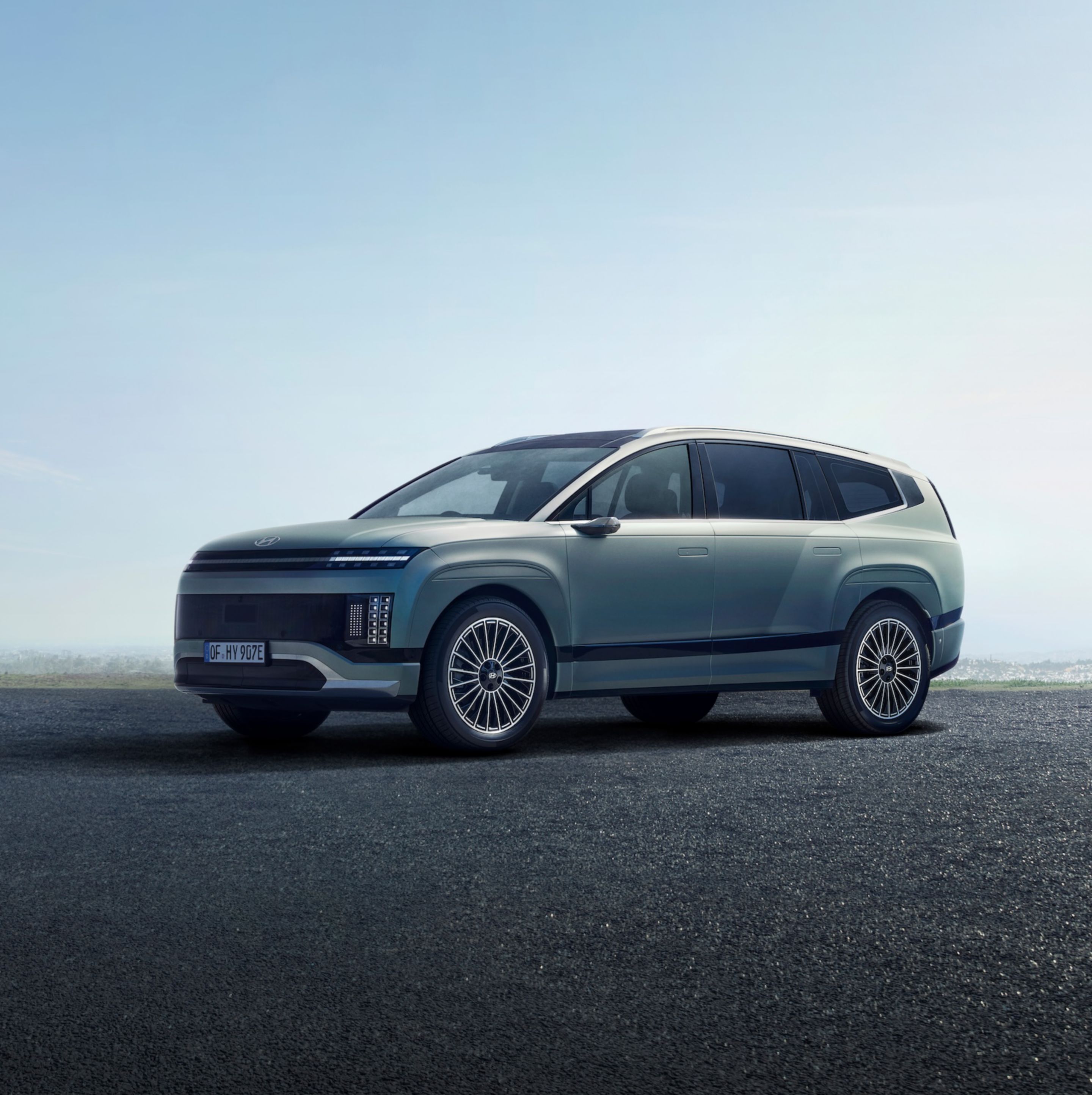 A Hyundai IONIQ 9 is parked on a paved surface under a clear sky, showcasing sleek design features like narrow headlights and large wheels.