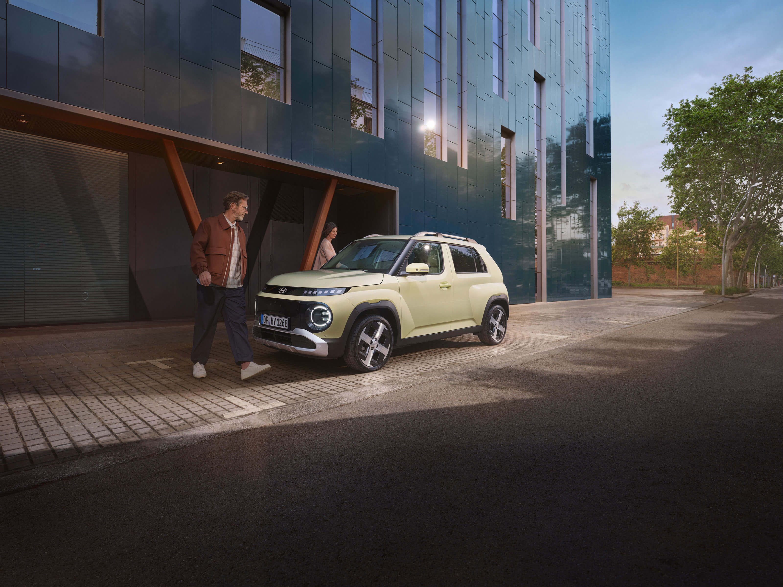Hyundai INSTER parked on a city sidewalk while two people walk past a modern building.