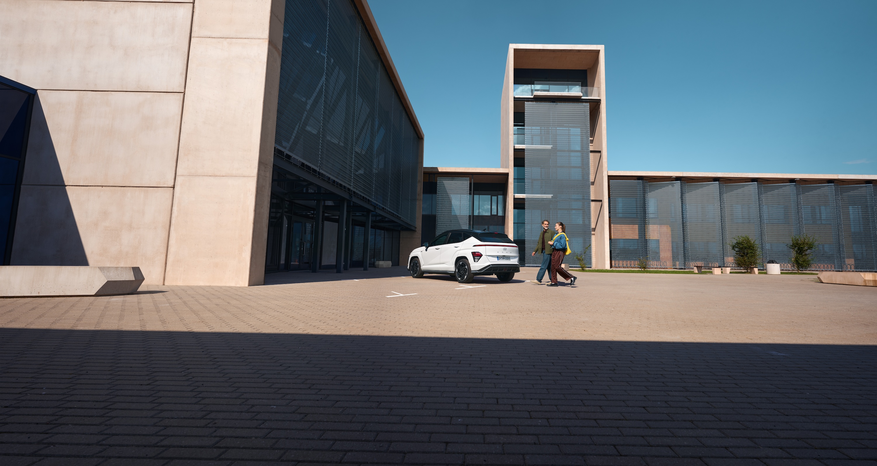 A white Hyundai KONA is parked on a paved lot next to modern buildings, as a man and a woman walk past under a clear blue sky.