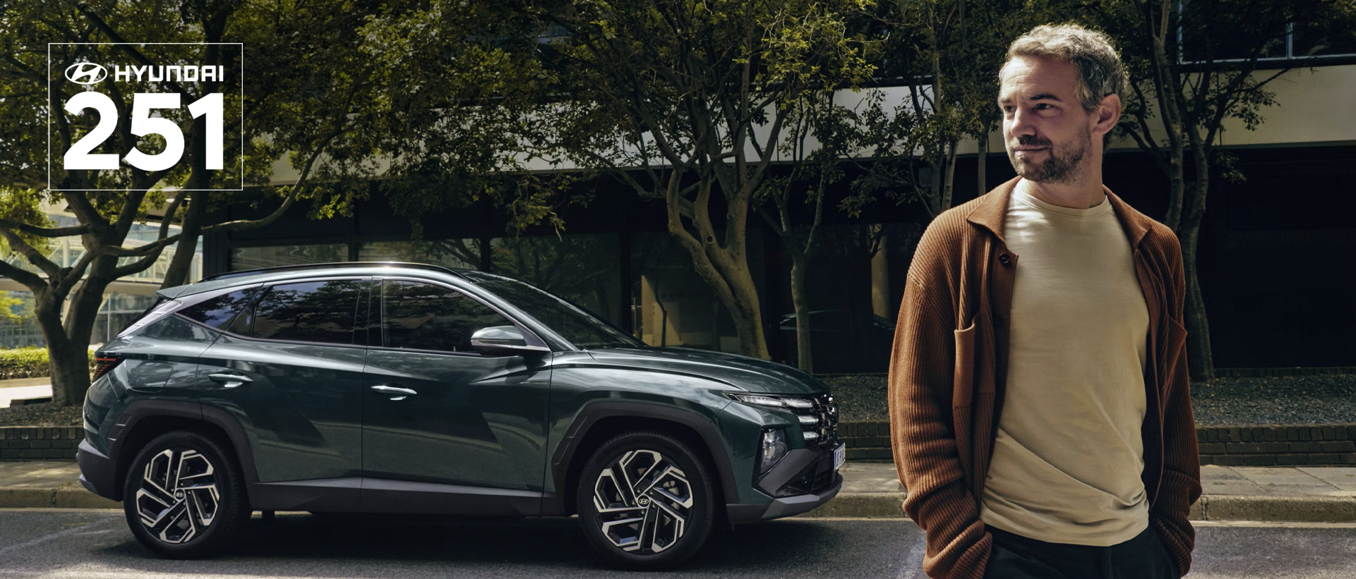 A man walking near a Hyundai TUCSON Hybrid parked by some trees.