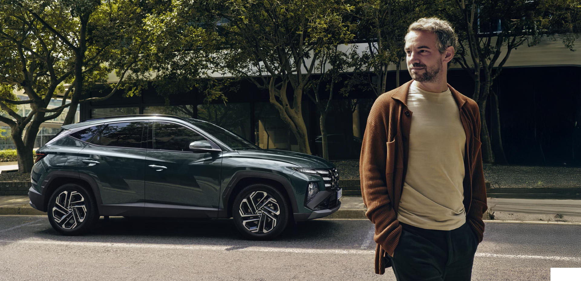 A man walking near a Hyundai TUCSON Hybrid parked by some trees.