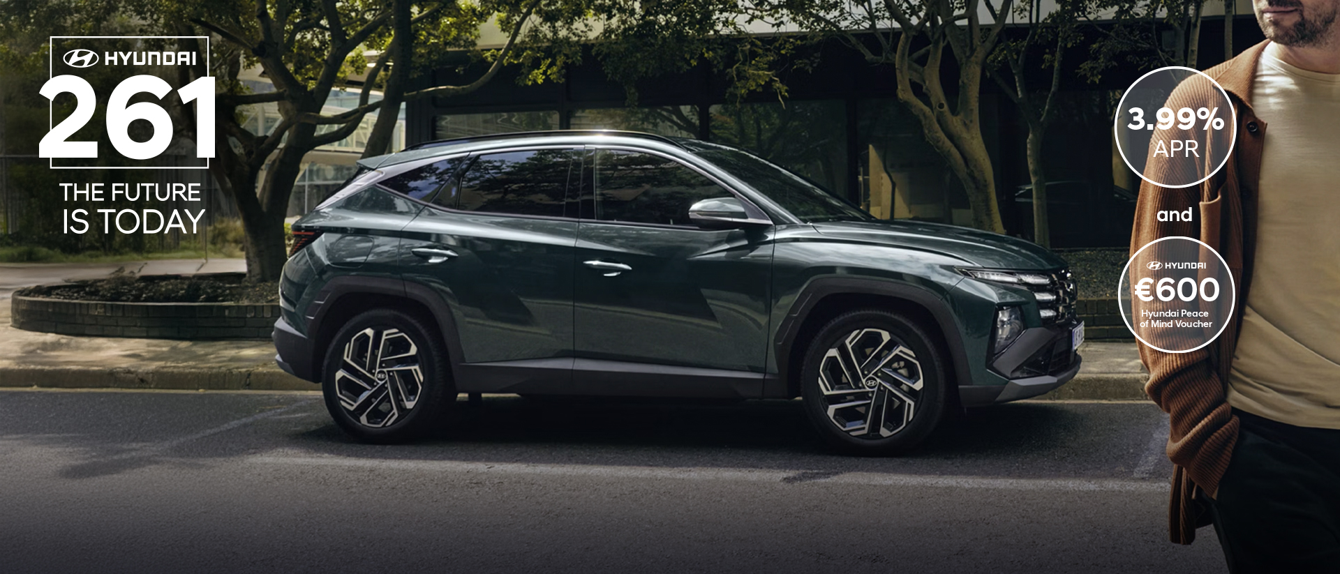 A man walking near a Hyundai TUCSON Hybrid parked by some trees.